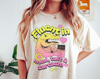 May include: A light beige t-shirt with a graphic of a cartoon alligator wearing a pink and yellow gradient shirt and holding a chocolate bar. The text "Fluent in mand, tact & sarcasm" is printed below the alligator. The shirt is labeled "Ivory".
