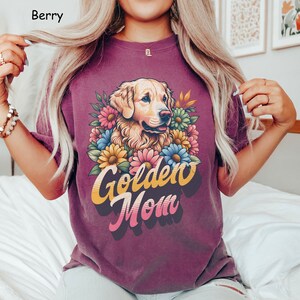 Puede incluir: Una camiseta morada con un perro golden retriever que lleva una corona de flores. El texto "Golden Mom" est&aacute; impreso en letras amarillas, naranjas y azules.