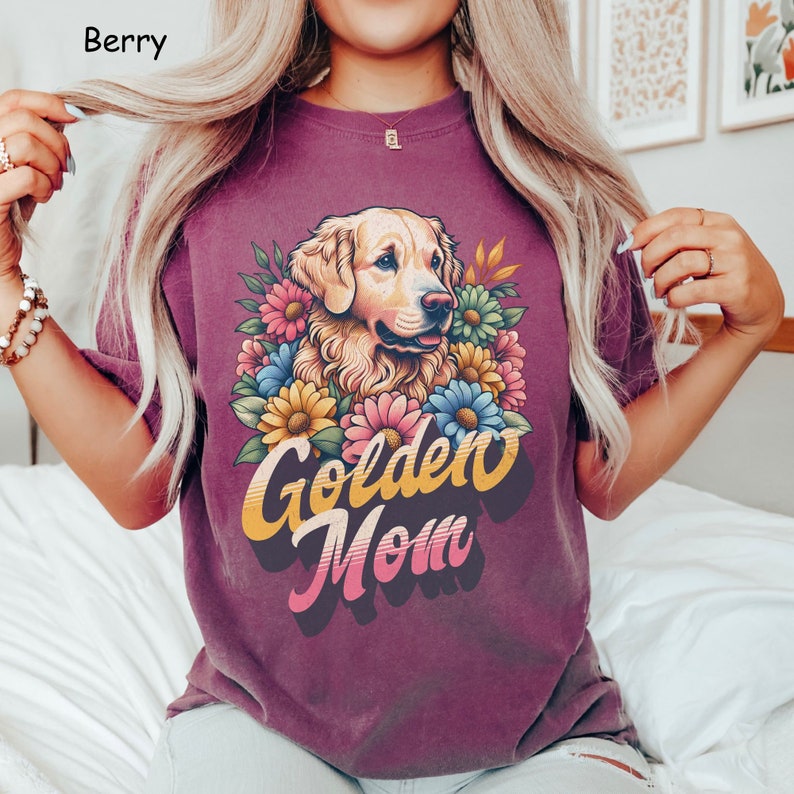 Puede incluir: Una camiseta morada con un perro golden retriever que lleva una corona de flores. El texto "Golden Mom" est&aacute; impreso en letras amarillas, naranjas y azules.
