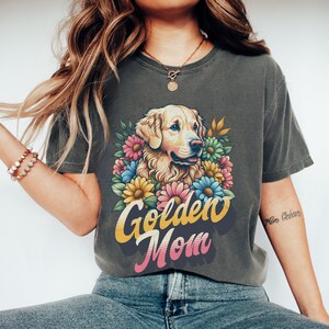 Puede incluir: Una camiseta gris oscuro con un gr&aacute;fico retro de un perro golden retriever rodeado de flores coloridas. El texto "Golden Mom" est&aacute; impreso en una fuente vintage debajo del perro.