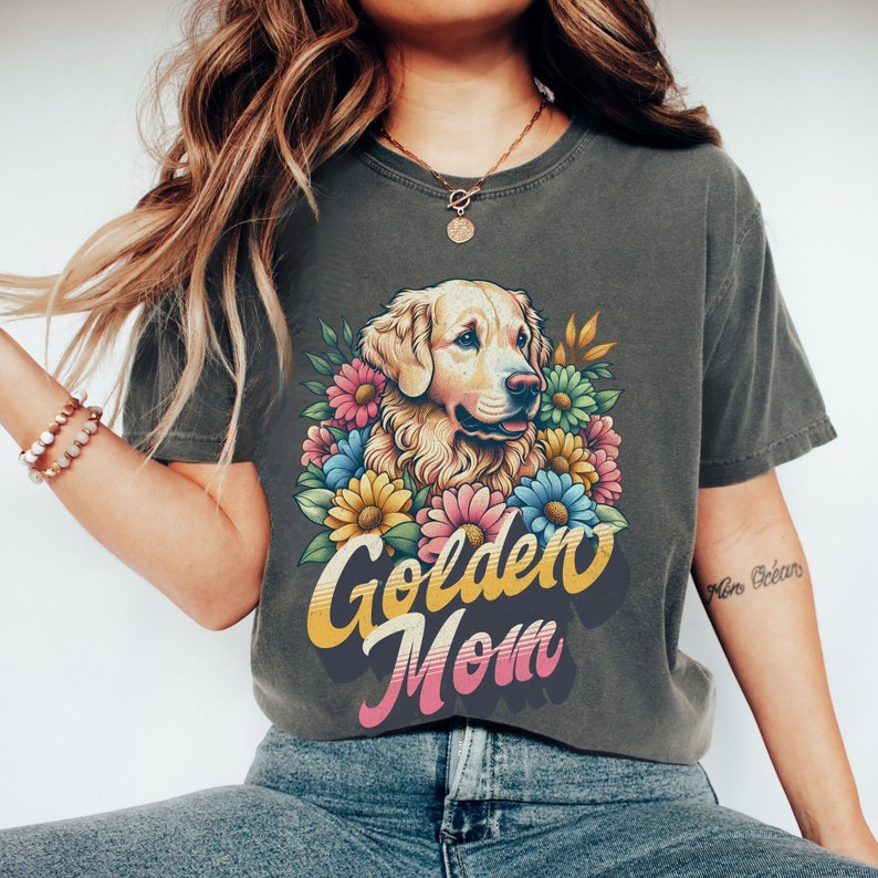 Puede incluir: Una camiseta gris oscuro con un gr&aacute;fico retro de un perro golden retriever rodeado de flores coloridas. El texto "Golden Mom" est&aacute; impreso en una fuente vintage debajo del perro.