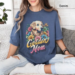 Puede incluir: Una mujer lleva una camiseta azul con una ilustraci&oacute;n de un perro golden retriever y el texto "Golden Mom" en una fuente retro. El perro est&aacute; rodeado de flores coloridas.
