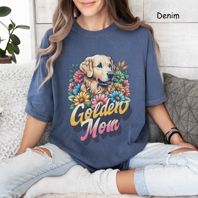 Puede incluir: Una mujer lleva una camiseta azul con una ilustraci&oacute;n de un perro golden retriever y el texto "Golden Mom" en una fuente retro. El perro est&aacute; rodeado de flores coloridas.