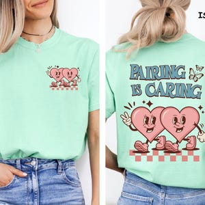 May include: A light green t-shirt featuring a retro design of two pink hearts with faces, hands, and feet. The front of the shirt has a small version of the design. The back of the shirt has the text "PAIRING IS CARING" with the same heart design.