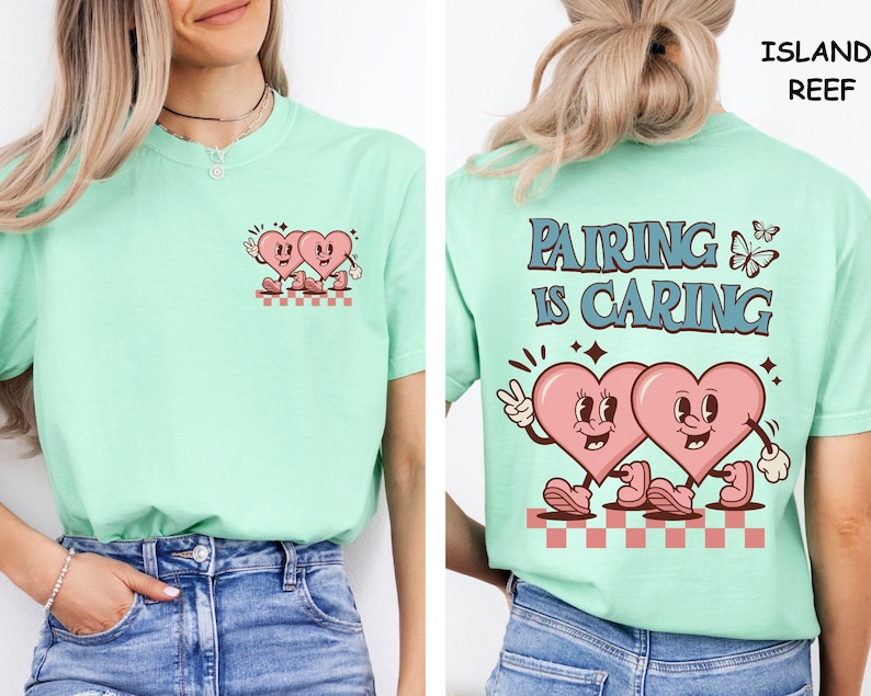 May include: A light green t-shirt featuring a retro design of two pink hearts with faces, hands, and feet. The front of the shirt has a small version of the design. The back of the shirt has the text "PAIRING IS CARING" with the same heart design.