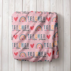 May include: Pink fabric with a repeating pattern of rainbows, hearts, and the text "TEACHER Mrs. Johnson".