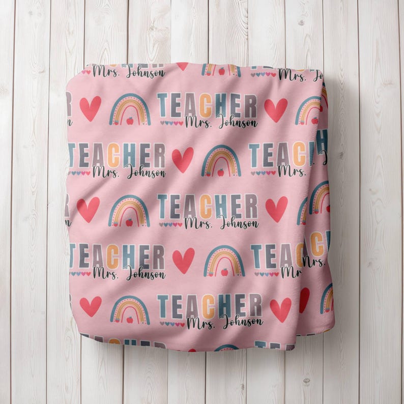 May include: Pink fabric with a repeating pattern of rainbows, hearts, and the text "TEACHER Mrs. Johnson".