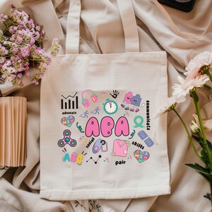 Könnte beinhalten: Cremefarbene Tragetasche mit einem farbenfrohen Design mit den Buchstaben "ABA" in Pink. Das Design enthält Symbole für Kopfhörer, eine Uhr, Puzzleteile und die Wörter "advocate", "social", "skills" und "communicate".