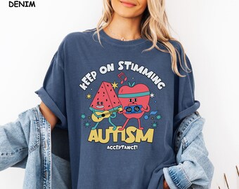 Può includere: Una maglietta blu navy con il testo "Keep on Stimming Autism Acceptance!" in giallo e bianco. Il design presenta un'anguria e una mela che suonano musica.