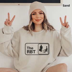 Op de afbeelding: Een beige sweatshirt met een zwarte afbeelding van een persoon die een koffer vasthoudt en een andere persoon die aan een bureau zit. De afbeelding zegt "The RBT" erboven.