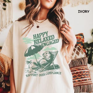 Puede incluir: Camiseta de color marfil con un gráfico de un castor relajándose en una silla de playa bajo una sombrilla. El texto dice "Happy Relaxed Engaged" y "Rapport Over Compliance".