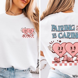 May include: White sweatshirt featuring a retro design with two pink cartoon hearts. The front has a small heart graphic, while the back displays the text "Pairing is Caring" with a butterfly and more hearts. The design is in a vintage style.