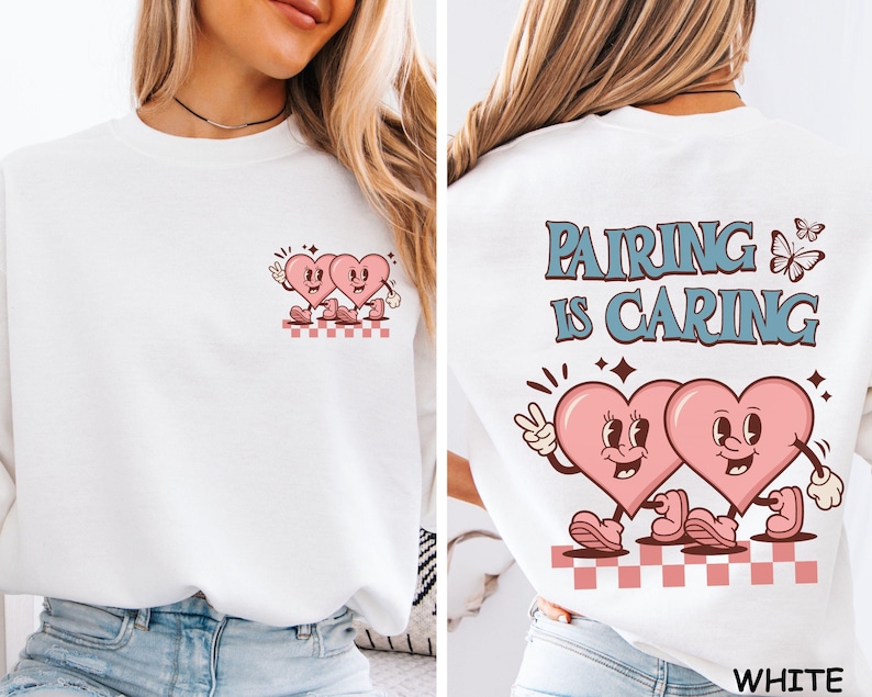 May include: White sweatshirt featuring a retro design with two pink cartoon hearts. The front has a small heart graphic, while the back displays the text "Pairing is Caring" with a butterfly and more hearts. The design is in a vintage style.