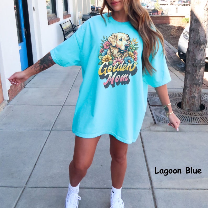 Puede incluir: Una camiseta de gran tama&ntilde;o de color azul claro con un gr&aacute;fico de un perro golden retriever con flores y el texto "Golden Mom". La camiseta se muestra en una modelo que lleva zapatillas blancas. El texto "Lagoon Blue" es visible en la esquina inferior derecha de la imagen.