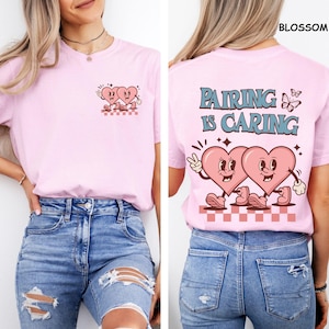 May include: Light pink t-shirt with a retro design featuring two cartoon hearts. The front has a small graphic of the hearts, while the back says "Pairing is Caring" with a larger heart graphic. The shirt is made of soft material.