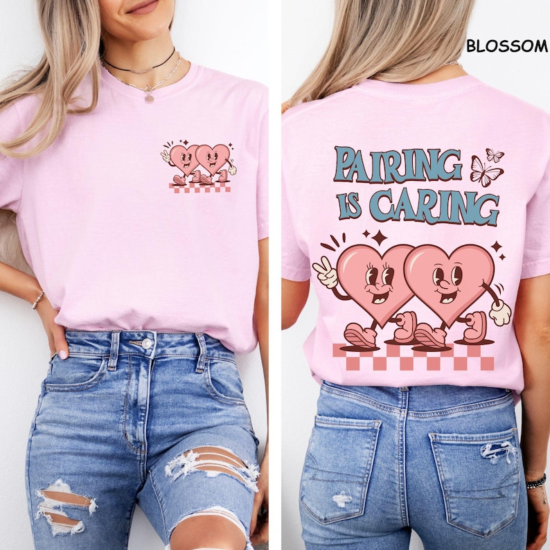 May include: Light pink t-shirt with a retro design featuring two cartoon hearts. The front has a small graphic of the hearts, while the back says "Pairing is Caring" with a larger heart graphic. The shirt is made of soft material.