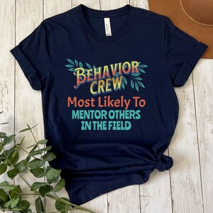 May include: Navy blue t-shirt with a colorful graphic that says "Behavior Crew Most Likely To Mentor Others In The Field".