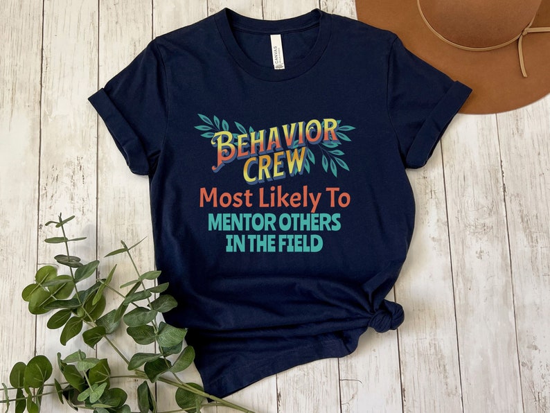 May include: Navy blue t-shirt with a colorful graphic that says "Behavior Crew Most Likely To Mentor Others In The Field".
