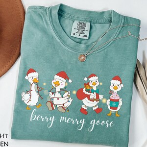 以下が含まれることがあります： ライトグリーンのComfort Colors Tシャツ。お祝いのガチョウのデザインが特徴です。グラフィックには、サンタ帽をかぶり、クリスマスをテーマにしたアイテムを持っている4羽のガチョウが含まれています。「berry merry goose」の文字がガチョウの下に印刷されています。