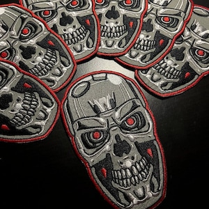 Puede incluir: Seis parches de calavera grises y plateados con contornos rojos. Las calaveras tienen ojos y dientes rojos. Los parches están dispuestos en semicírculo.