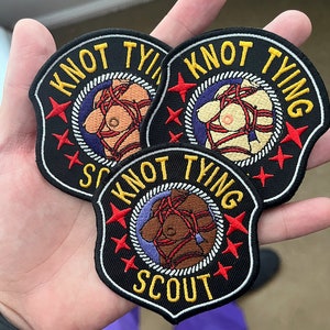 Puede incluir: Tres parches bordados con fondo negro, costuras blancas y texto amarillo. Los parches muestran una figura desnuda atada con una cuerda roja y el texto "Knot Tying Scout".