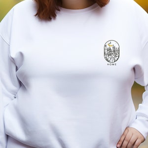 K&ouml;nnte beinhalten: Wei&szlig;er Pullover mit einem schwarzen und goldenen Grafikdruck eines Schlosses mit dem Wort "HOME" darunter.