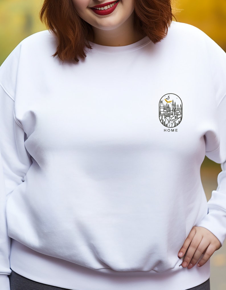K&ouml;nnte beinhalten: Wei&szlig;er Pullover mit einem schwarzen und goldenen Grafikdruck eines Schlosses mit dem Wort "HOME" darunter.