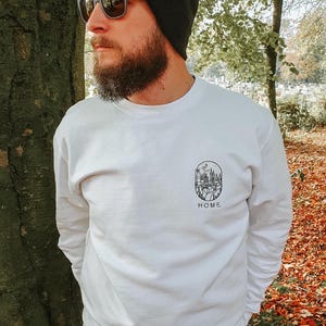 K&ouml;nnte beinhalten: Wei&szlig;es Sweatshirt mit einem schwarzen Aufdruck einer Stadtlandschaft mit einem dar&uuml;ber fliegenden Vogel. Der Aufdruck befindet sich in einem Kreis mit dem Wort "HOME" darunter.
