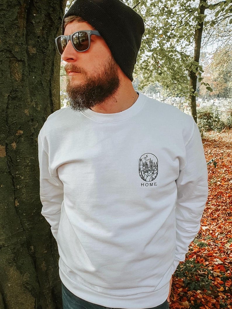K&ouml;nnte beinhalten: Wei&szlig;es Sweatshirt mit einem schwarzen Aufdruck einer Stadtlandschaft mit einem dar&uuml;ber fliegenden Vogel. Der Aufdruck befindet sich in einem Kreis mit dem Wort "HOME" darunter.