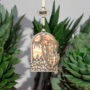 May include: A wooden ornament with a cutout design of a desert scene with a sun and cactus. The ornament is hanging from a string with a wooden tag that says "ROY".