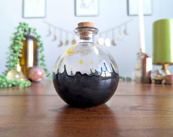 Puede incluir: Una botella de vidrio transparente con tapón de corcho, con una base negra y un diseño celestial de estrellas amarillas y una luna creciente. La botella está sobre una superficie de madera.