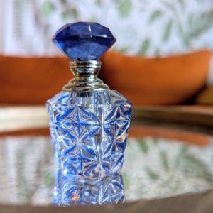May include: A clear glass perfume bottle with a faceted, diamond-shaped blue stopper. The bottle has a geometric design and a silver-colored neck. The bottle is sitting on a reflective surface.