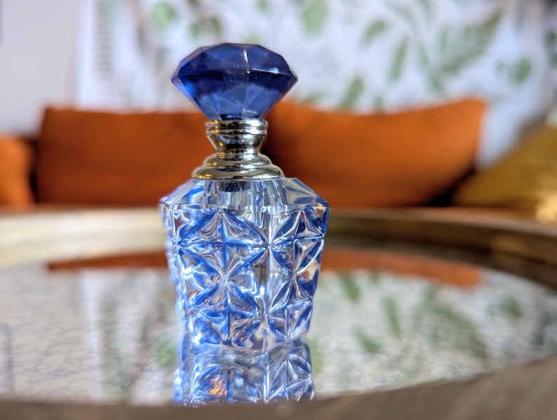 May include: A clear glass perfume bottle with a faceted, diamond-shaped blue stopper. The bottle has a geometric design and a silver-colored neck. The bottle is sitting on a reflective surface.