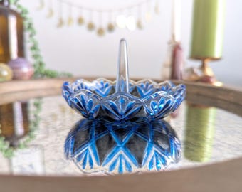 May include: A blue glass ring holder with a scalloped edge and a central handle. The ring holder has a geometric design and is reflecting on a mirrored surface. The background includes a candle and decorative items.