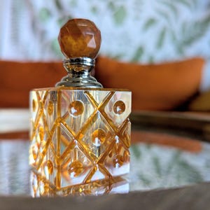 May include: A decorative amber-colored perfume bottle with a faceted, geometric design. The bottle has a square shape with a silver-colored neck and a round, amber-colored stopper. The bottle is sitting on a reflective surface.