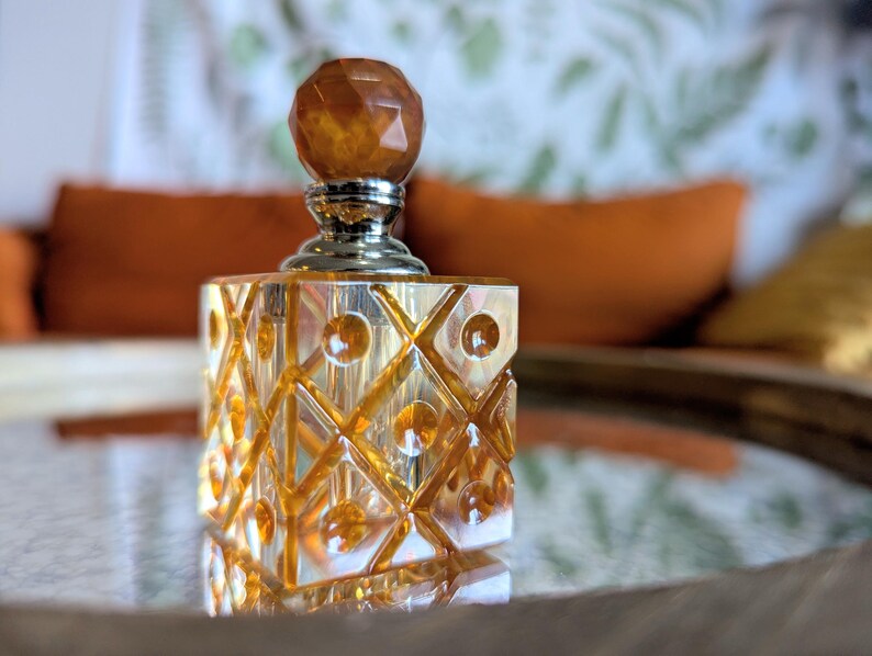 May include: A decorative amber-colored perfume bottle with a faceted, geometric design. The bottle has a square shape with a silver-colored neck and a round, amber-colored stopper. The bottle is sitting on a reflective surface.