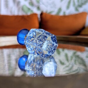 May include: A faceted, clear glass decorative object with a blue sphere attached. The object is reflected on a mirrored surface. The background includes orange pillows and a patterned fabric.