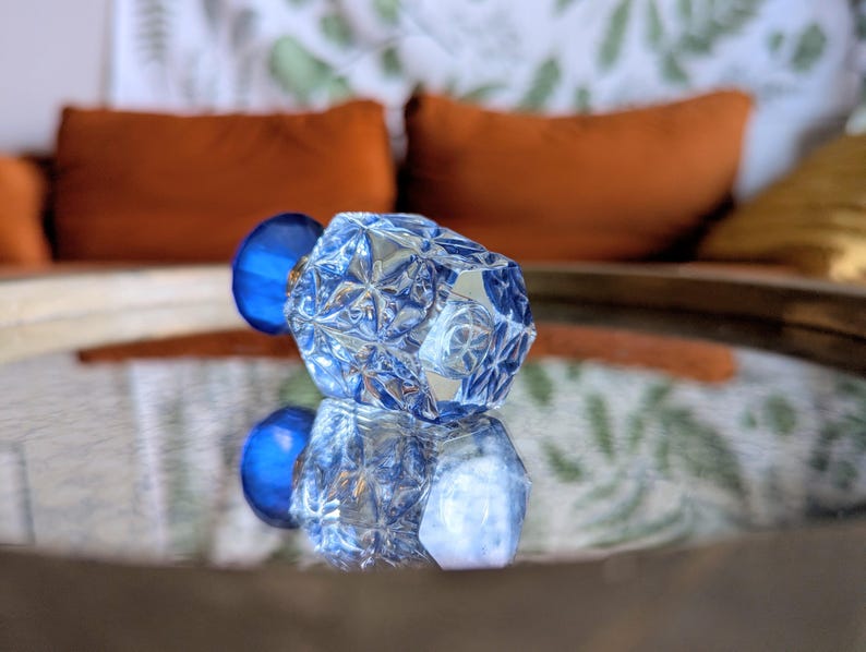May include: A faceted, clear glass decorative object with a blue sphere attached. The object is reflected on a mirrored surface. The background includes orange pillows and a patterned fabric.