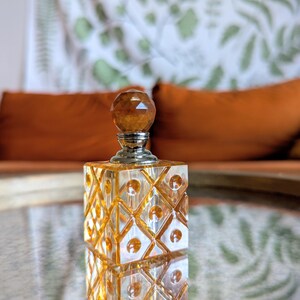 May include: A decorative perfume bottle with a faceted amber-colored stopper and a square, patterned body. The bottle sits on a reflective surface, with a blurred background featuring orange cushions and a patterned wall hanging.