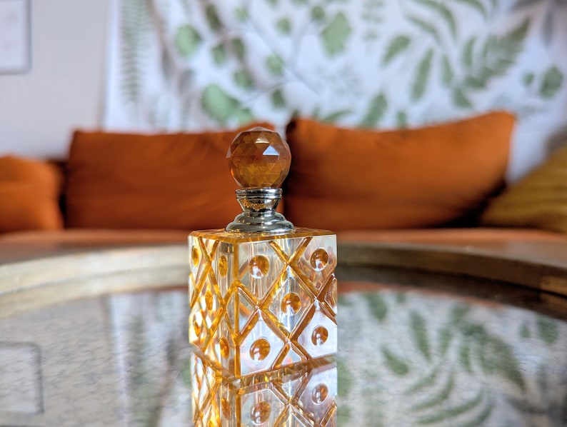 May include: A decorative perfume bottle with a faceted amber-colored stopper and a square, patterned body. The bottle sits on a reflective surface, with a blurred background featuring orange cushions and a patterned wall hanging.