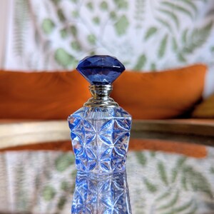 May include: A blue glass perfume bottle with a faceted, diamond-shaped stopper. The bottle has a silver-colored neck and a geometric pattern. The background includes orange and yellow pillows and a patterned curtain.