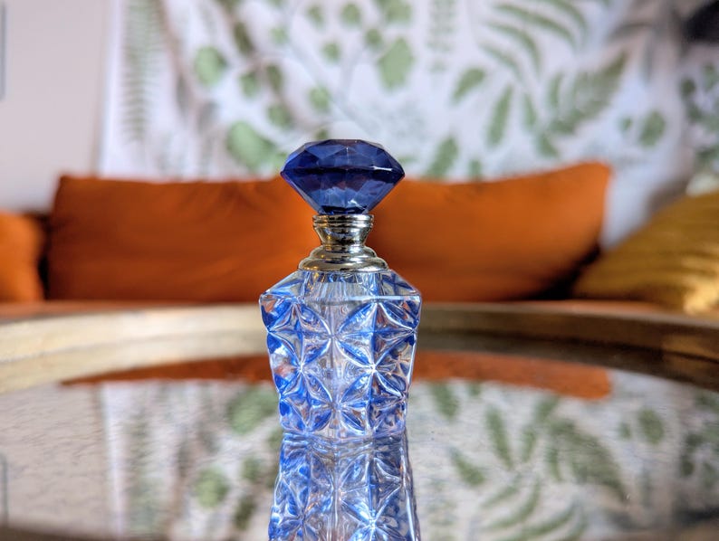 May include: A blue glass perfume bottle with a faceted, diamond-shaped stopper. The bottle has a silver-colored neck and a geometric pattern. The background includes orange and yellow pillows and a patterned curtain.