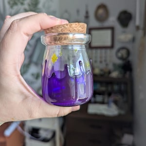 Könnte beinhalten: Ein kleines, klares Glasgefäß mit einem Korken, gefüllt mit einer leuchtend violetten Flüssigkeit. Das Glas ist mit gelben sternförmigen Formen und violetten Tropfen verziert. Das Glas wird in einer Hand gehalten.