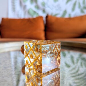 May include: A clear, square glass object with a geometric pattern and gold accents. The object is reflected on a mirrored surface. The background features orange pillows and a patterned wall.