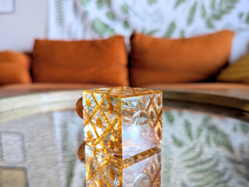 May include: A clear, square glass object with a geometric pattern and gold accents. The object is reflected on a mirrored surface. The background features orange pillows and a patterned wall.