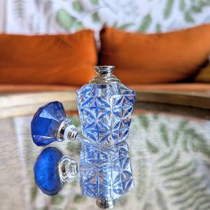 May include: A clear, faceted glass perfume bottle with a blue stopper. The bottle and stopper are reflecting on a mirrored surface. The bottle has a silver-colored top. The background includes orange pillows and a patterned wall.