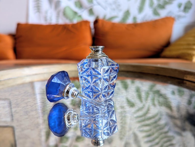 May include: A clear, faceted glass perfume bottle with a blue stopper. The bottle and stopper are reflecting on a mirrored surface. The bottle has a silver-colored top. The background includes orange pillows and a patterned wall.