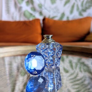May include: A clear, faceted glass perfume bottle with a silver-colored top and a large, blue, gem-like stopper. The bottle and stopper are on a reflective surface, with orange pillows in the background.