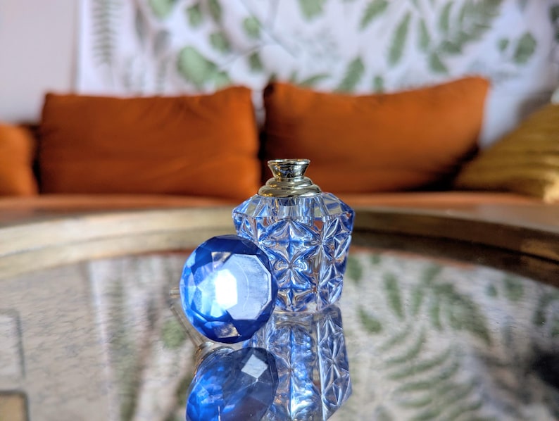 May include: A clear, faceted glass perfume bottle with a silver-colored top and a large, blue, gem-like stopper. The bottle and stopper are on a reflective surface, with orange pillows in the background.