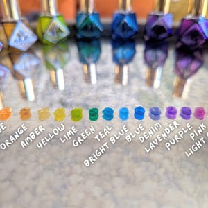 May include: A collection of colorful nail polish bottles with gold caps, reflecting on a surface. Below, a row of matching color swatches labeled with names like "Red," "Orange," "Green," and "Purple."